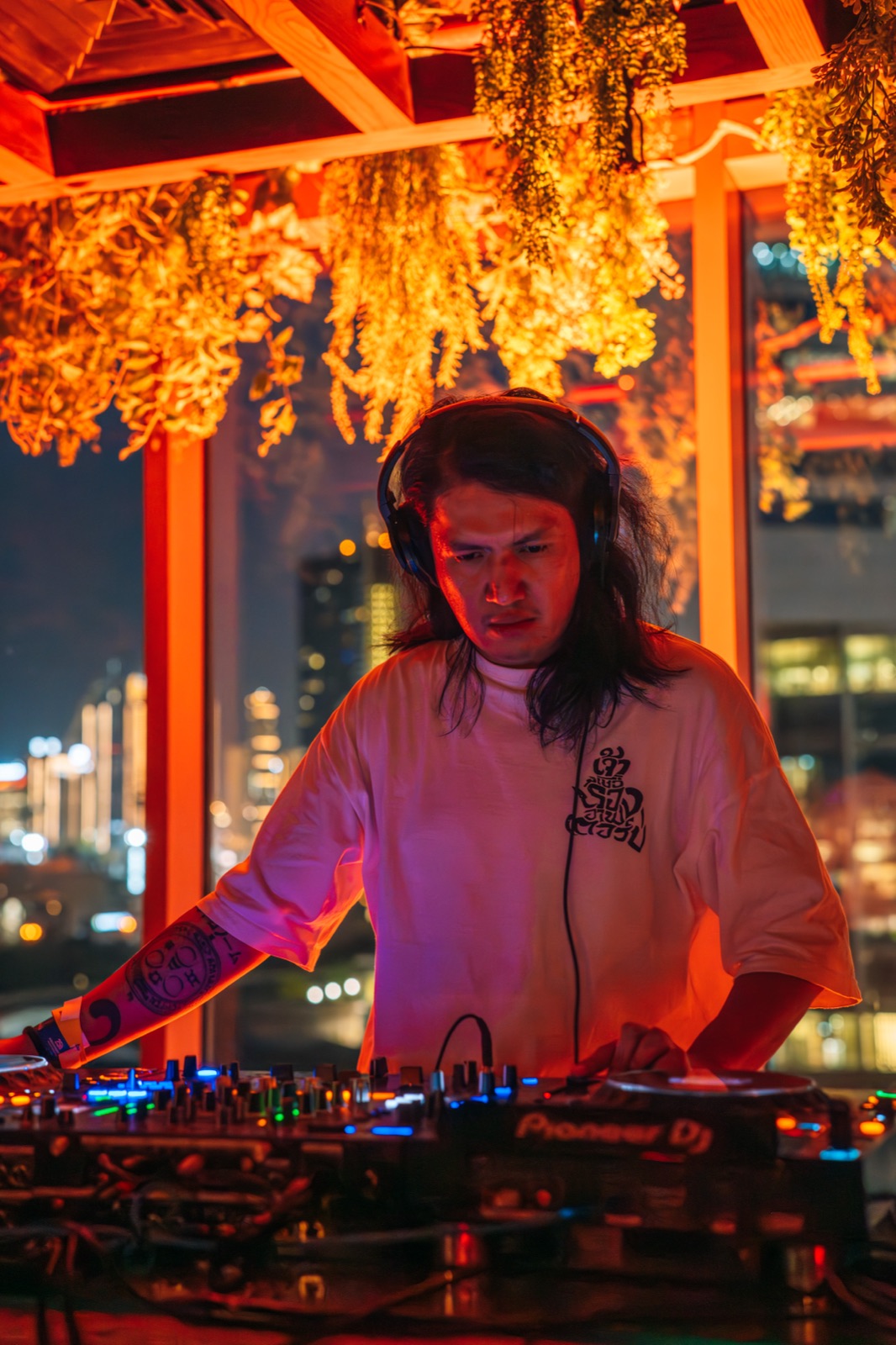 A DJ with long hair mixing at night, with city lights behind.