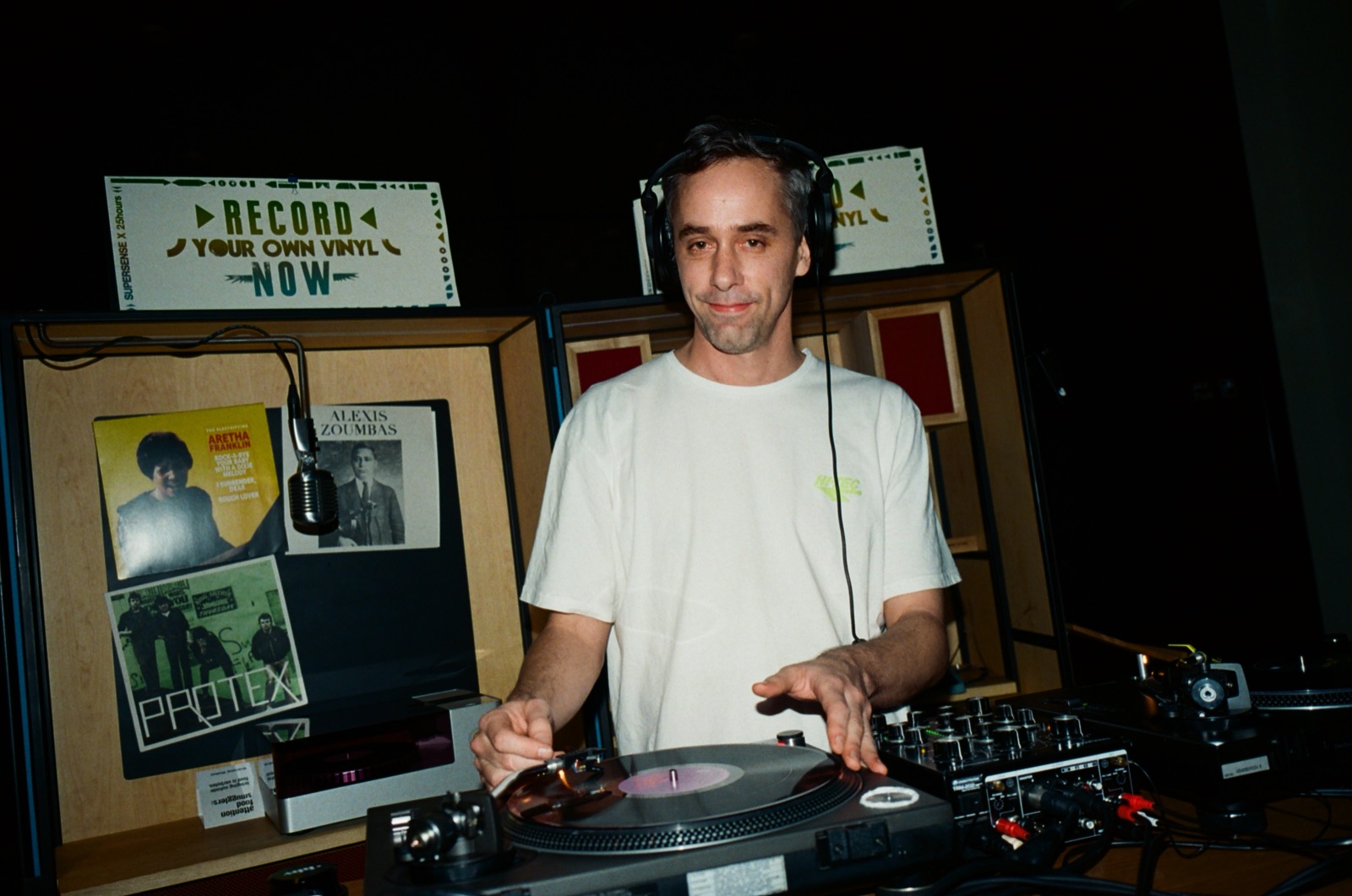 A DJ in a white T-shirt playing records in front of a Record Store Now sign.