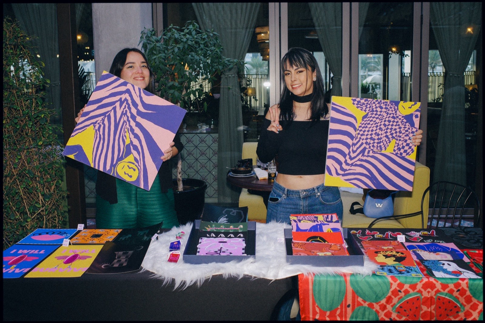 Two women holding up purple and yellow artwork at a stall.