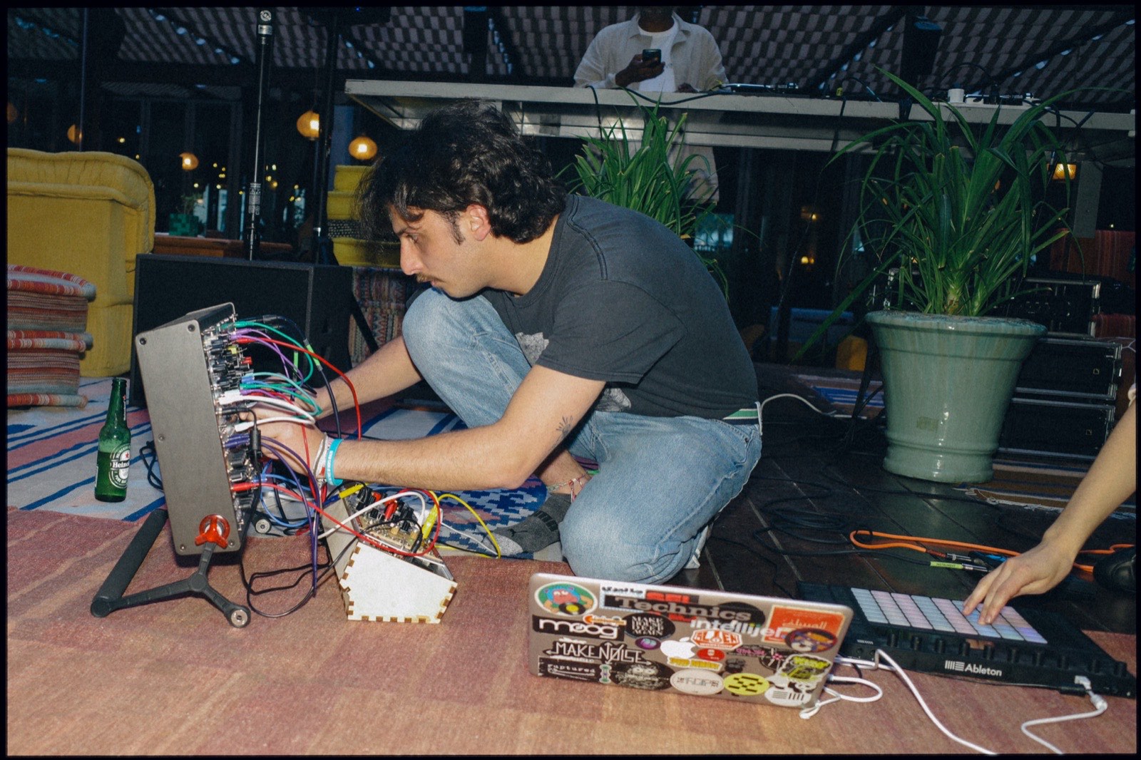 A musician crouched on a rug among synthesisers and cables.