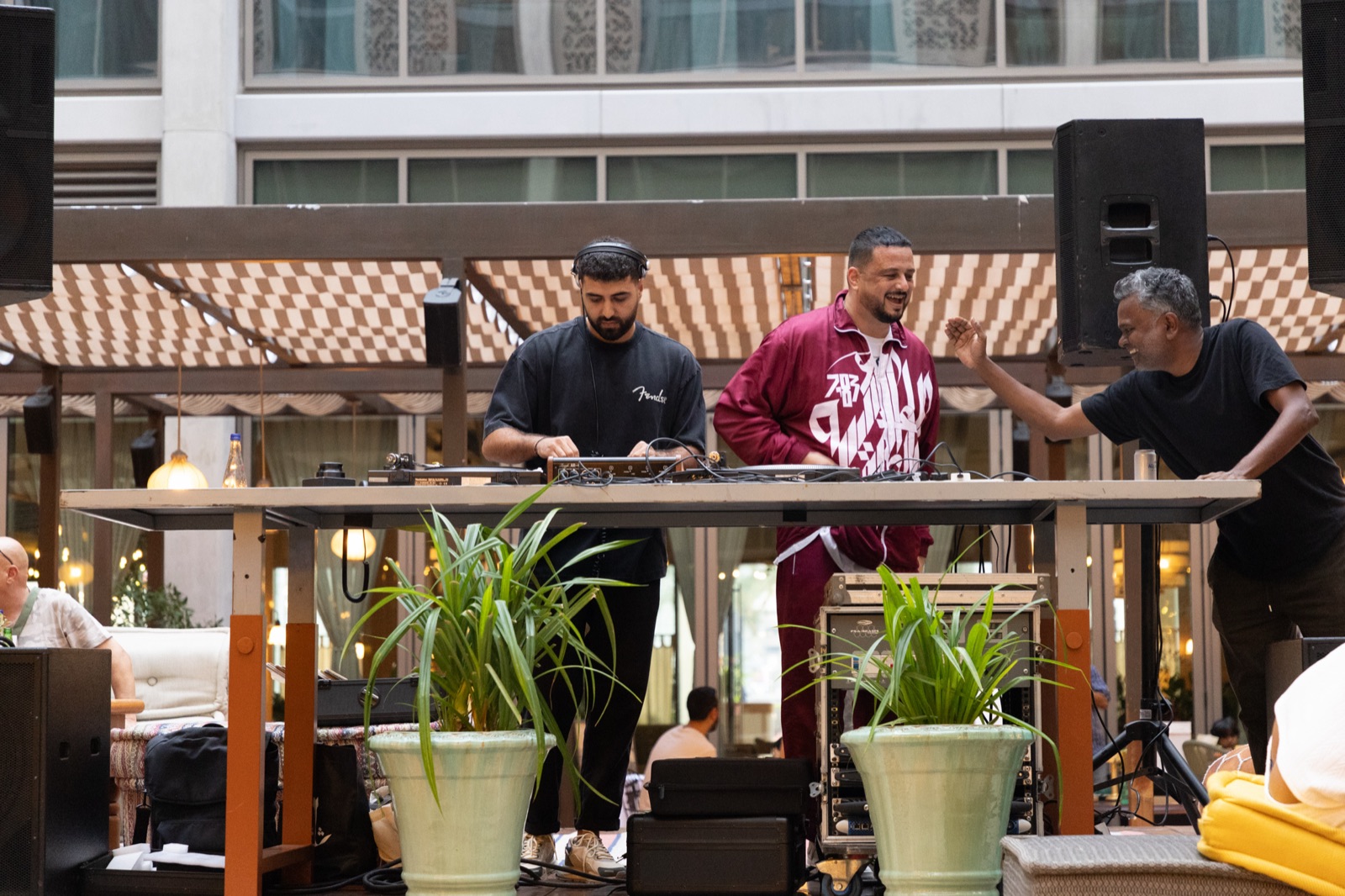 DJs playing at a long table on a sunny terrace with potted plants.