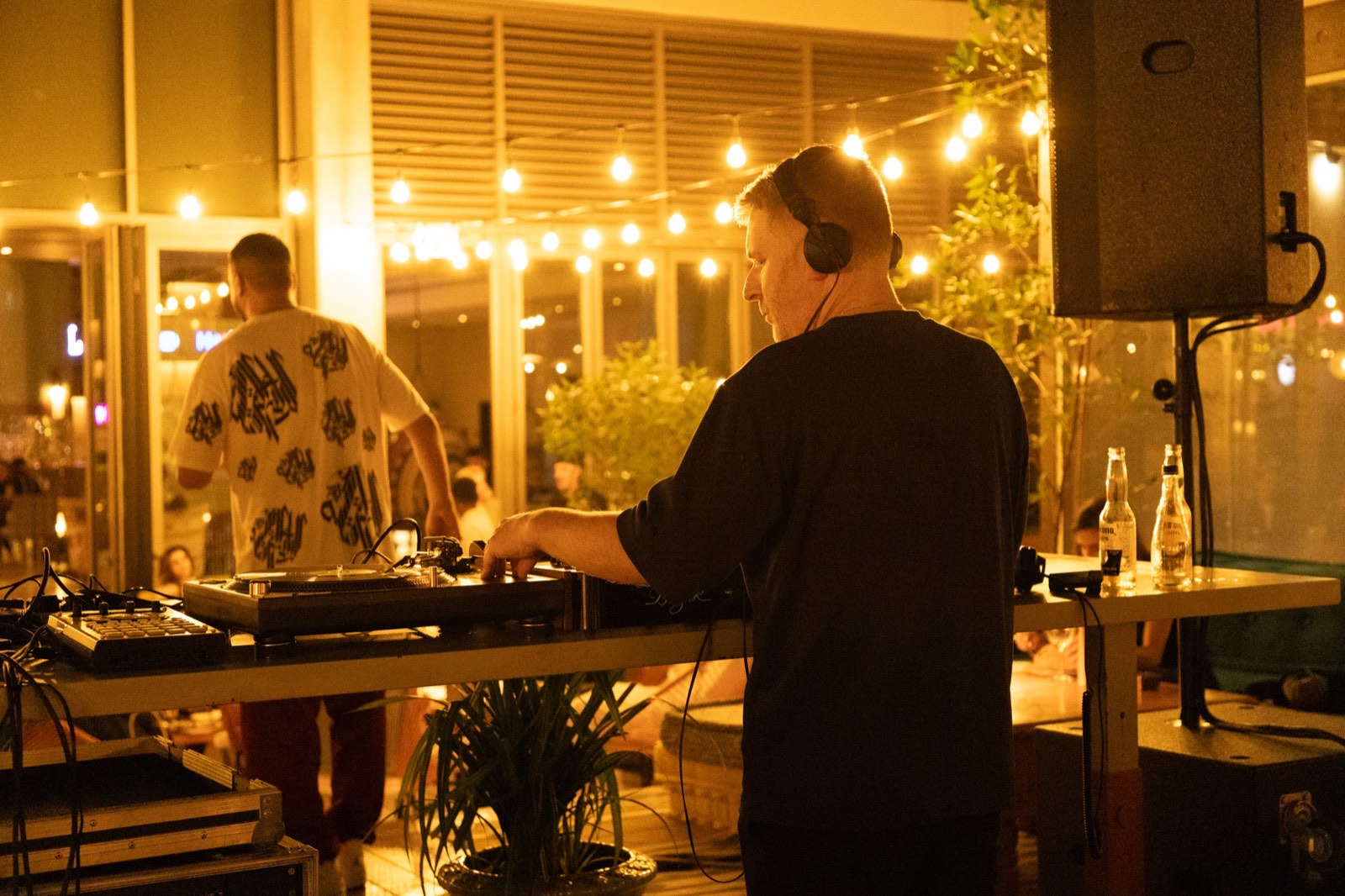 A DJ in headphones at a booth lit by strings of warm lights.