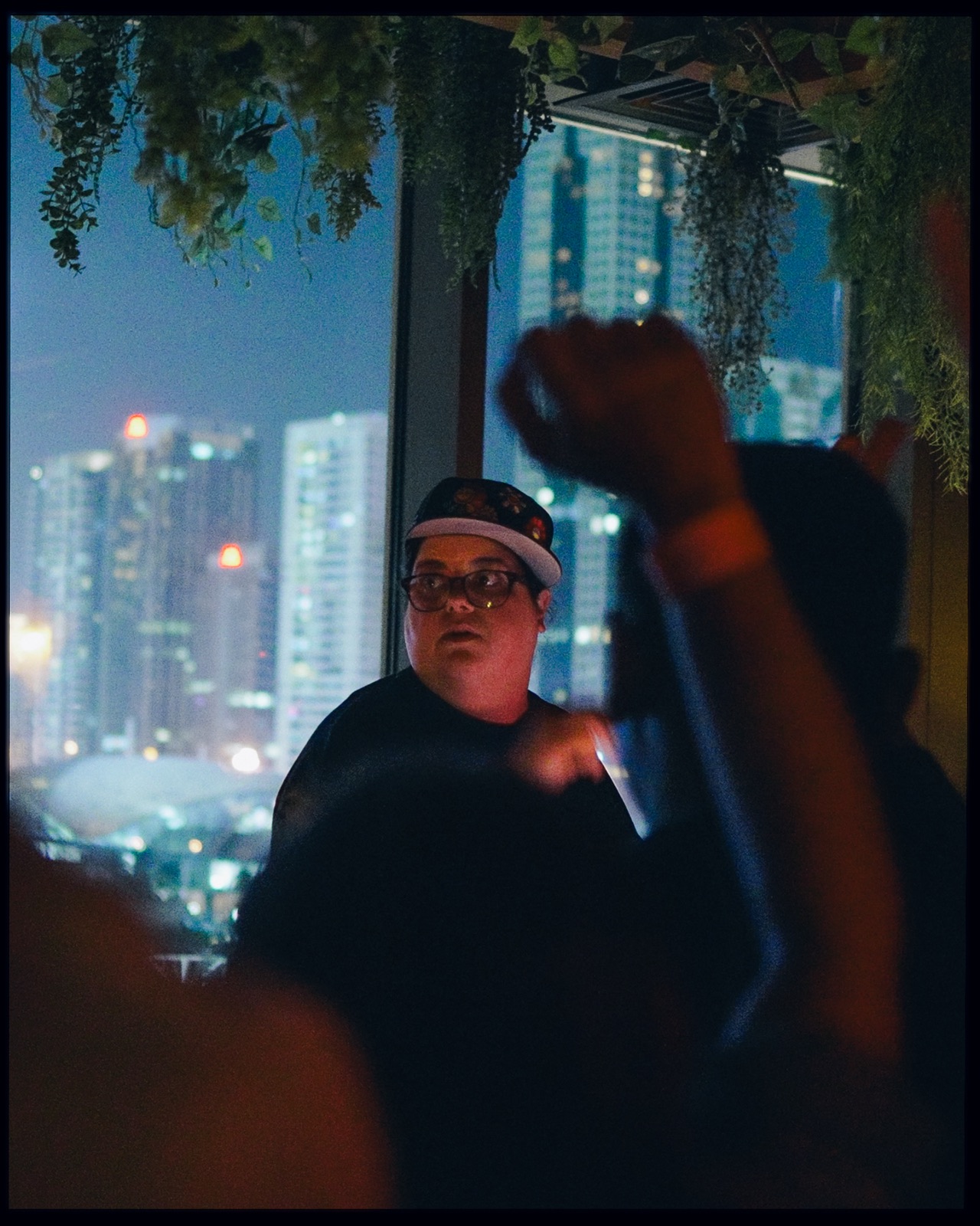 A DJ in a cap by a window, the city at night behind and a fist raised in the foreground.