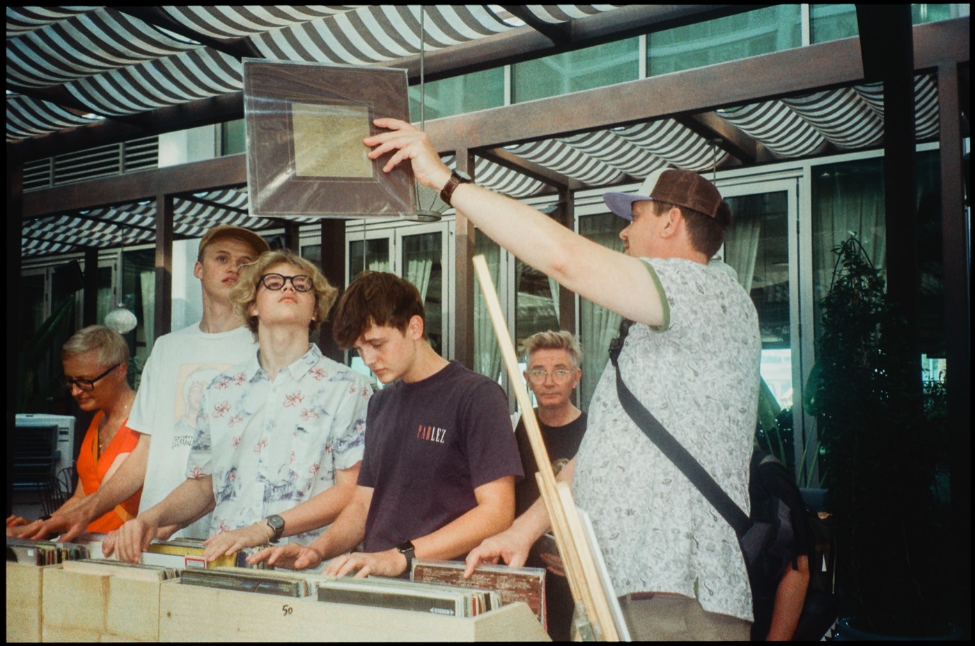 People digging through crates of records under a striped awning, one holding a record up to look at it.