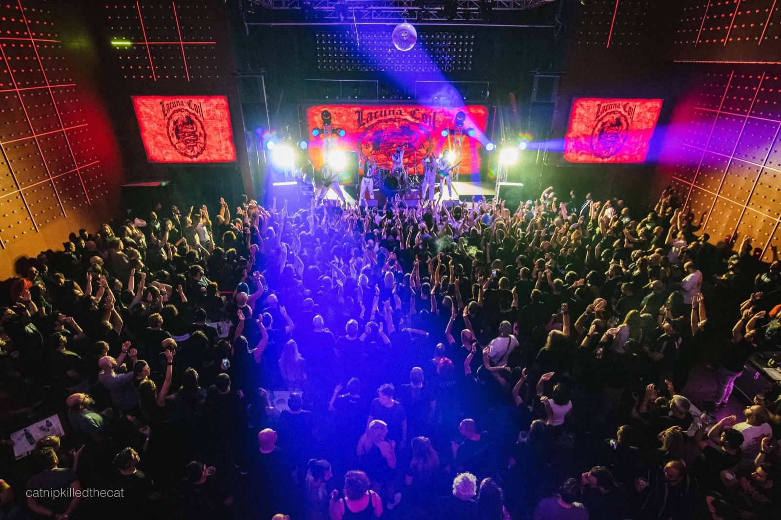 A packed venue seen from above, with blue and red stage lights over a crowd facing the band.